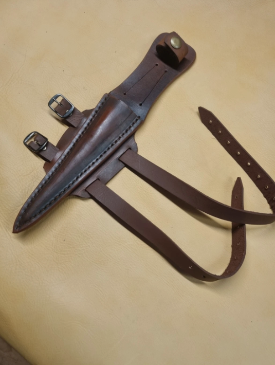 A dark brown leather knife sheath with a matching adjustable strap and buckle, laid flat on a beige textured surface.