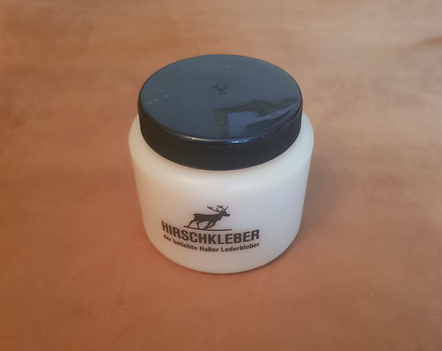 Hirschkleber paste glue tub adhesive for leather and fabric. Cream tub with black plastic lid displayed n tan leather background