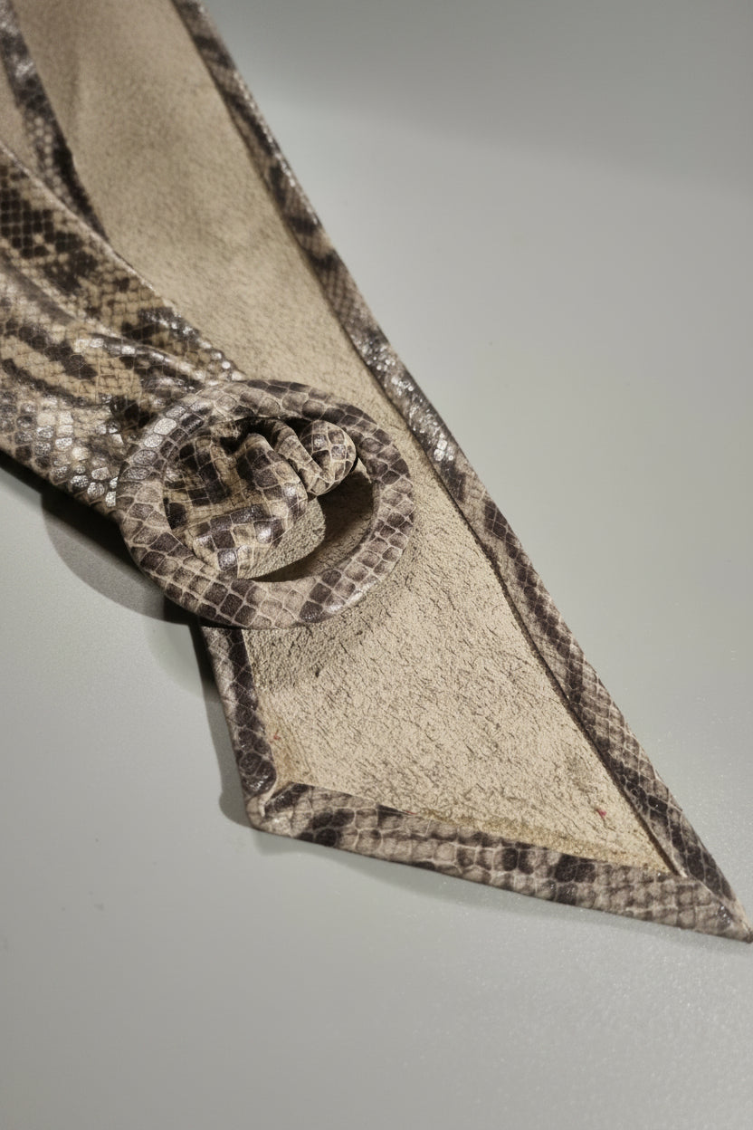 A close-up of a snakeskin-patterned belt with a large round buckle, laid diagonally on a neutral gray surface.