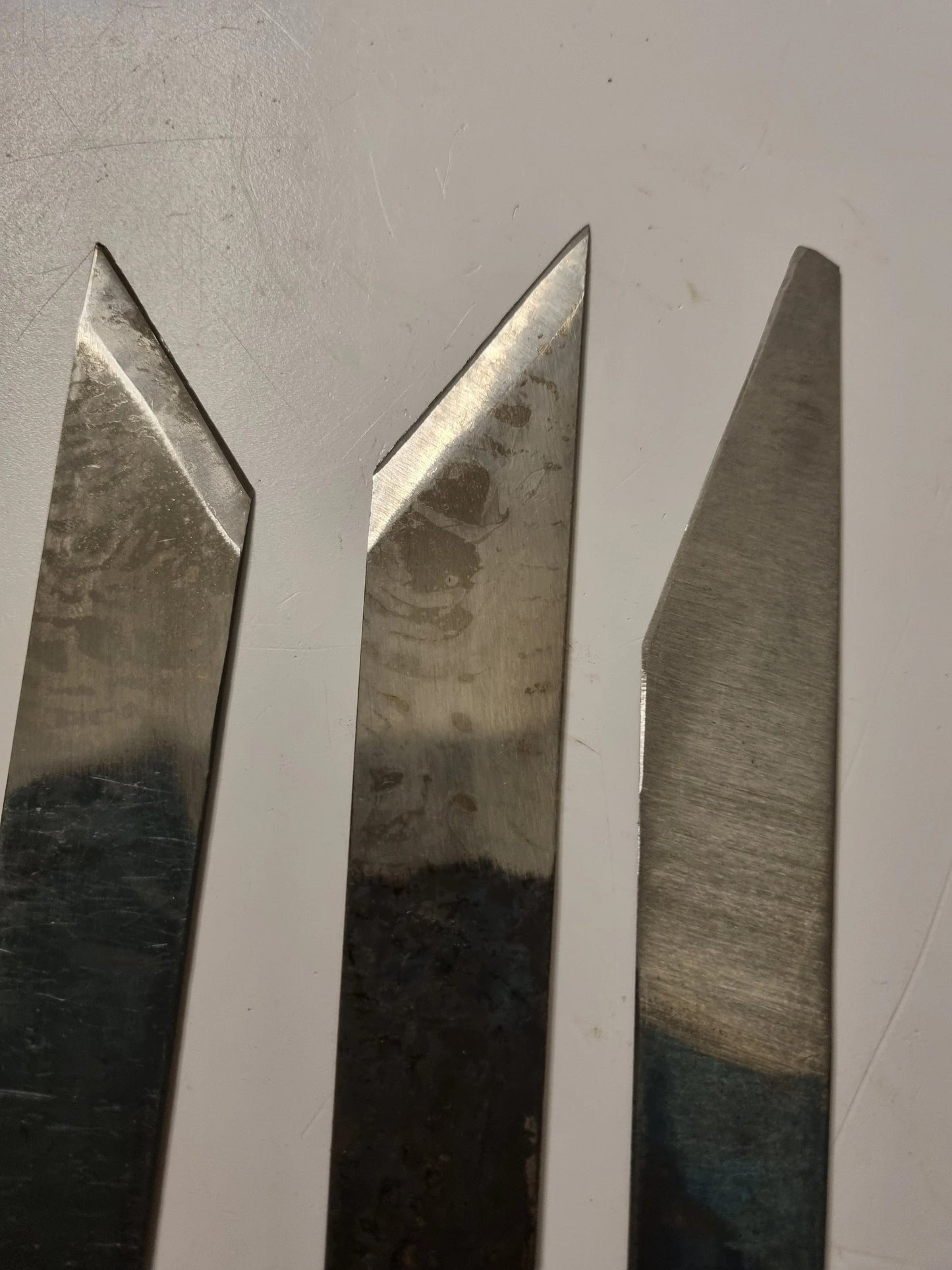 Three sharpened metal knife blades or cutting tools standing vertically against a plain light background.