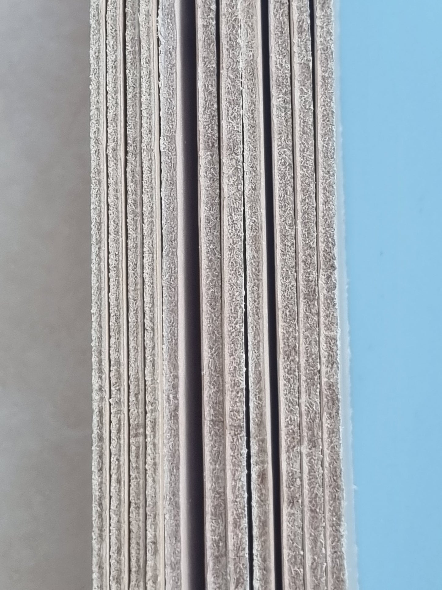 A close-up of a stack of textured gray panels or boards standing vertically, with a light blue background visible on the right side.