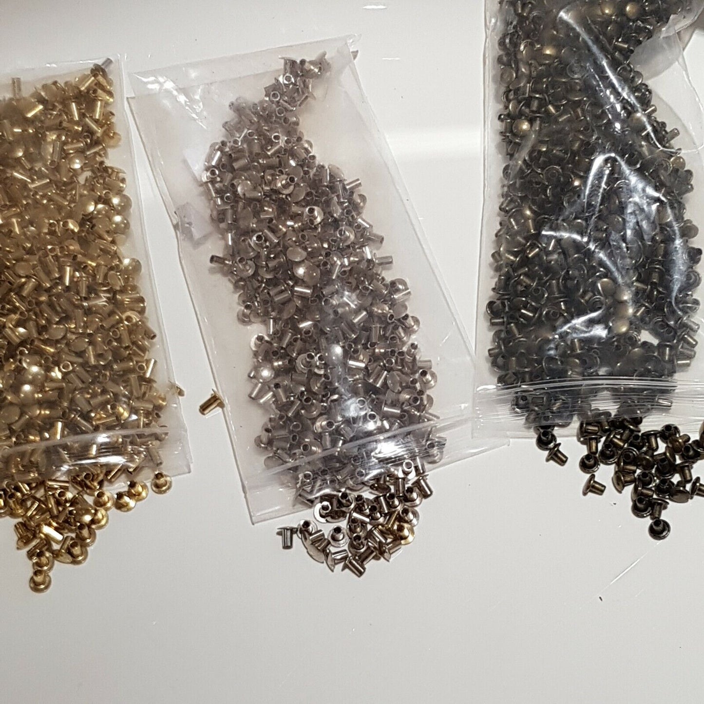 Three bags of semi tubular rivets in gold, brass, and nickel colors, displayed on a white surface.