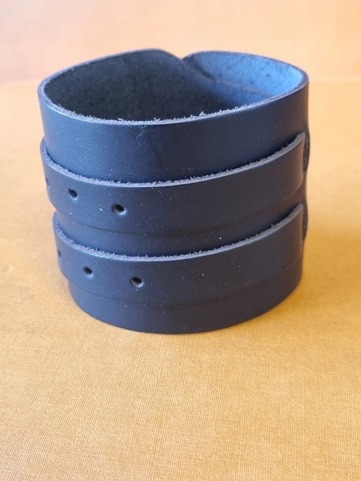 A black leather cuff bracelet with 2strap bands and pre-drilled holes, displayed on a textured tan surface.