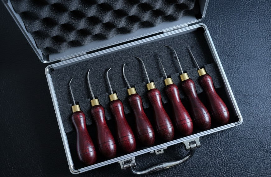 A set of eight leatherworking hand stitch awls with dark reddish-brown wooden handles and gold metal ferrules, arranged inside an open silver aluminum carrying case with a black foam insert, photographed on a dark textured surface.