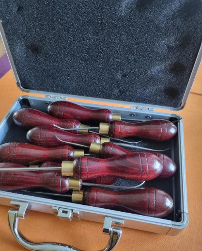 A set of hand sewing awls with reddish-brown wooden handles and brass ferrules collars, displayed inside an open silver metal case with a black foam insert.