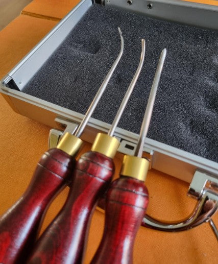 Three curved metal tools with reddish-brown wooden handles protected with brass ferrules, displayed in front of an open gray foam-lined case on an orange surface.