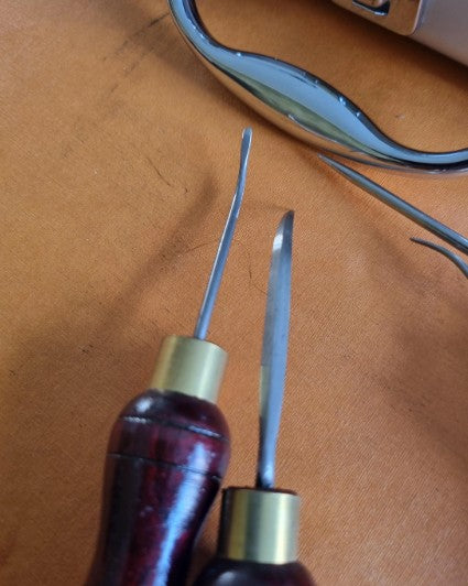 Two hand sewing welt awls with dark reddish-brown wooden handles and brass ferrules, shown on a tan leather-like surface. A curved metallic object is partially visible in the background.