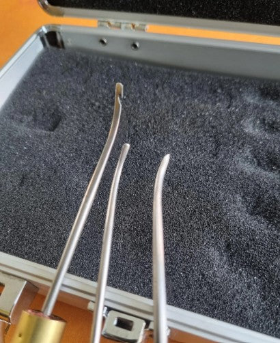 Three curved metal awls resting in a black foam-lined hard case with a silver metal frame.