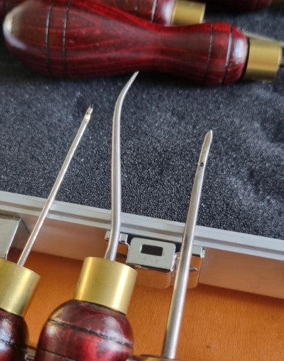 Close-up of a set of hand sewing awls with reddish-brown wooden handles and brass ferrules, resting on a dark textured surface next to a silver metal case.