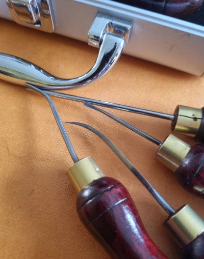 Close-up of a leatherworking hand sewing awls set with curved metal hooks and sharp pointed tips, featuring dark reddish-brown wooden handles with brass ferrules collars, resting on a tan leather surface.