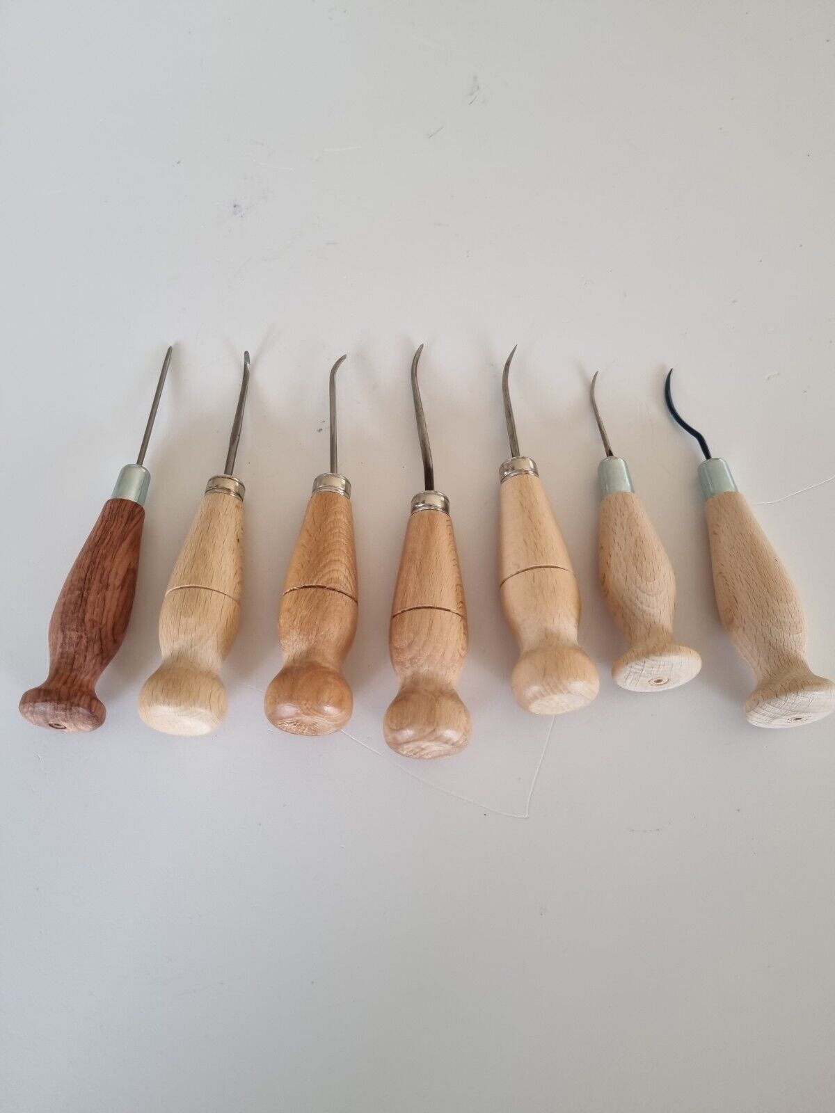 7 types of leather craft hand stitching  awls shaped straight, curved and hooked. All 7 awls inserted into light colour birch wood handles on a white background