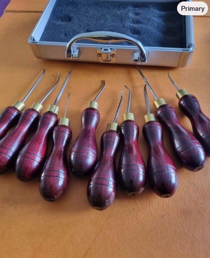 A set of 10 hand awls for leathercraft work sewing tools with dark reddish-brown wooden handles and brass metal ferrules, arranged on a wooden surface with an open silver carrying case behind them.