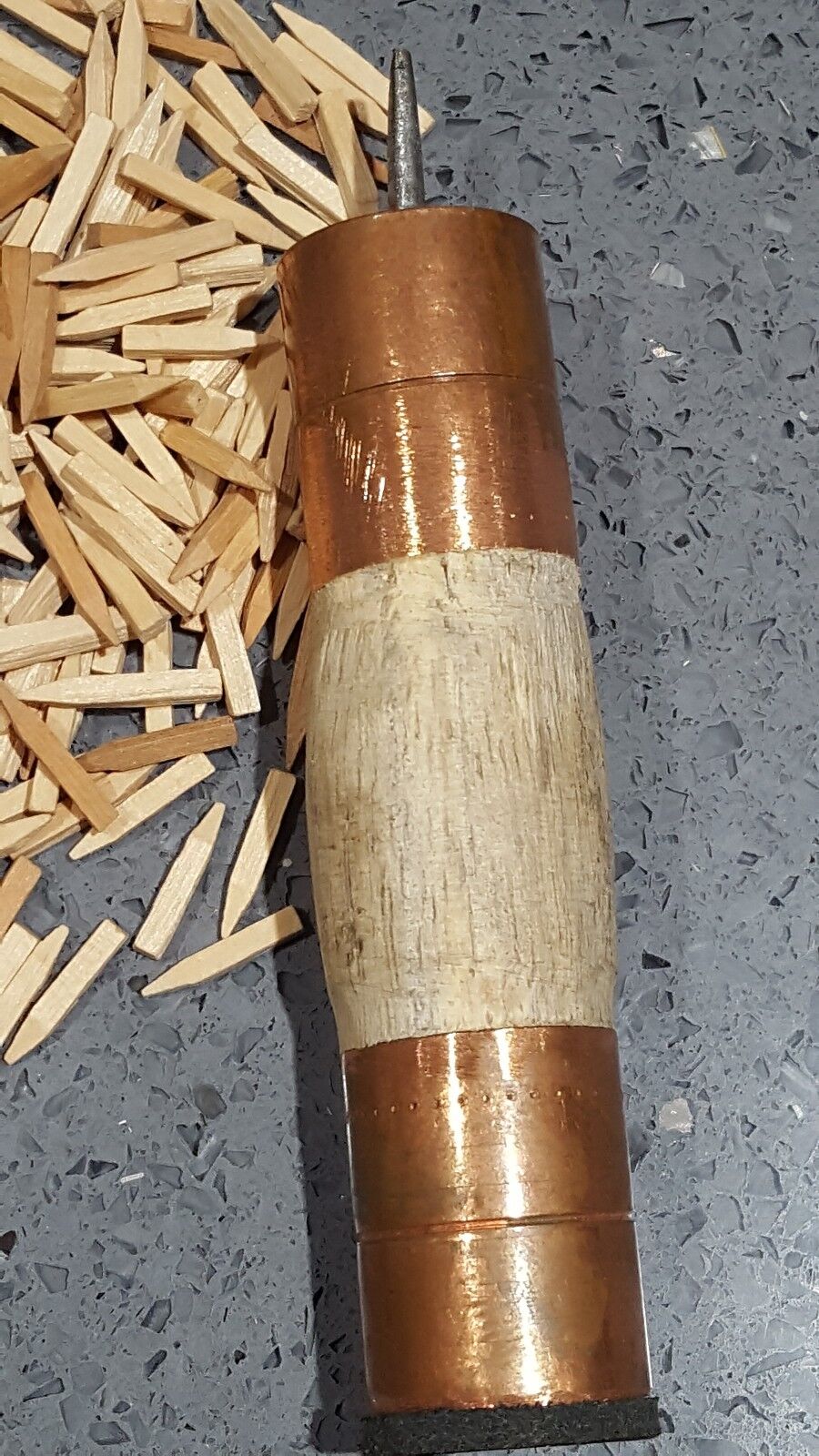 A wooden handle with brass ferrules and sharp awl for driving wooden pegs or nails into leather shown next to heap of wooden pegs against grey background