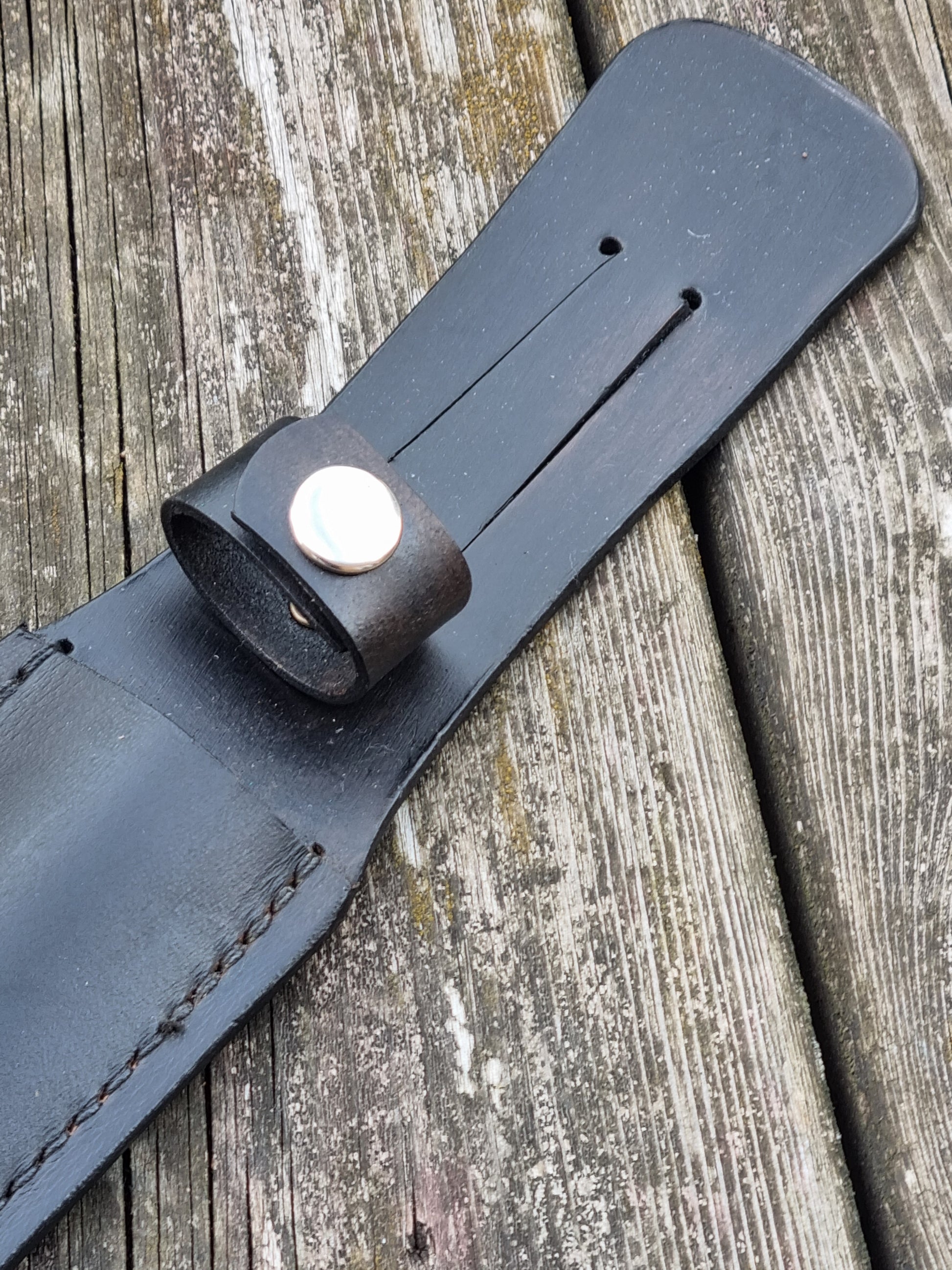 Close-up of a black leather sheath strap with belt slot and a handle snap loop  on a wooden surface