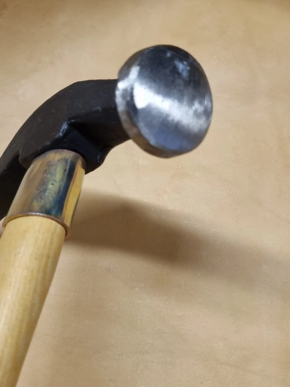 A close-up of a hammer with a polished steel ball-shaped head, a black metal neck, and a light wood handle, set against a plain tan background.