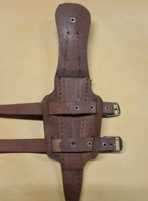 A brown leather dagger sheath turned over to show two adjustable straps and metal buckles, laid flat on a tan textured background.