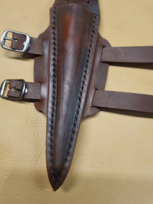 A dark brown leather knife sheath with a pointed tip, stitched along the edges, and attached to two adjustable leather straps with silver metal buckles, laid flat on a beige textured surface.