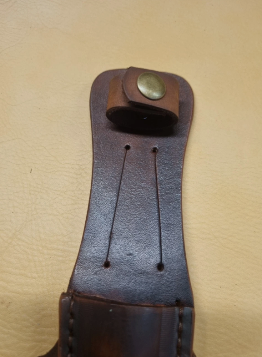 A close-up of a dark brown leather sheath  handle loop with brass snap button, shown against a light beige textured background.