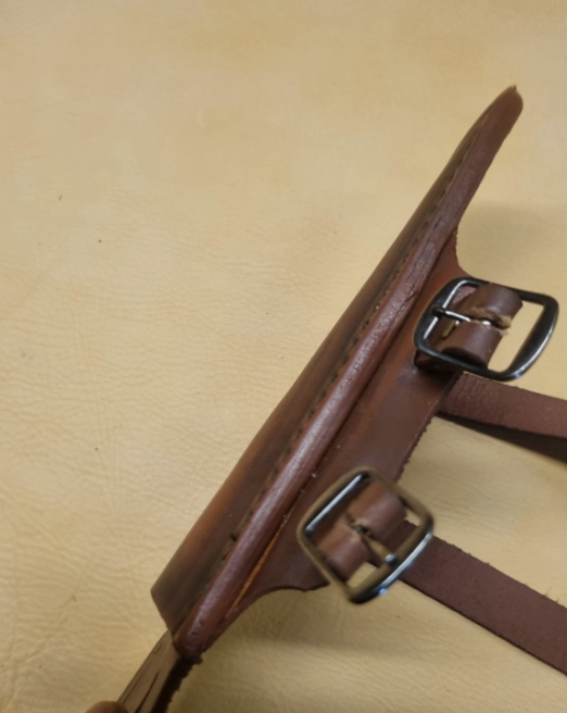 A close-up of a brown leather strap component with two dark metal buckles, laid on a tan textured surface.