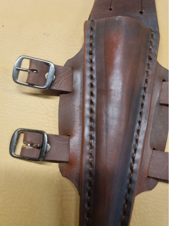 A close-up of a brown leather dagger  sheath with visible stitching, metal buckles, and straps laid on a tan surface.