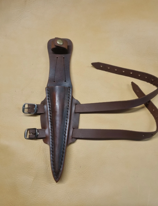 A dark brown leather sheath with a long strap and metal buckles, laid flat on a light tan leather background.