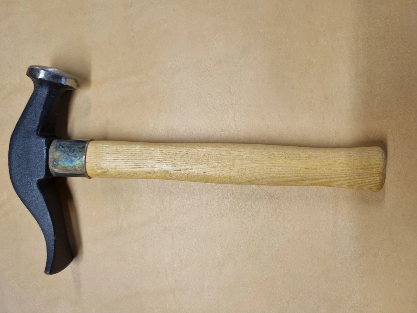A French hammer with a black metal head and a light-coloured wooden handle, placed horizontally on a plain tan surface.