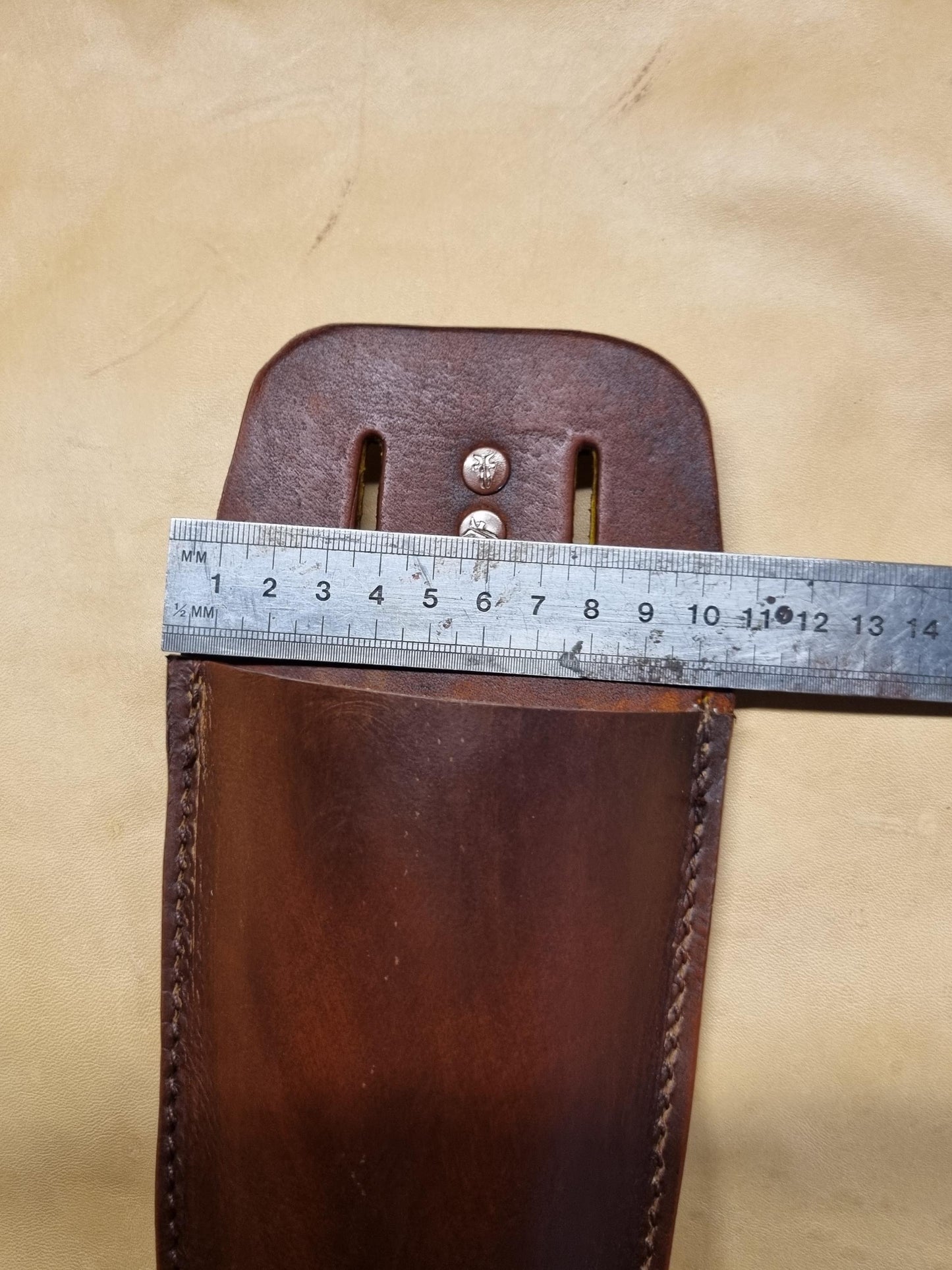 A close-up of a brown leather secateurs holster with a metal ruler laid across the top to show its width, set against a beige fabric background.