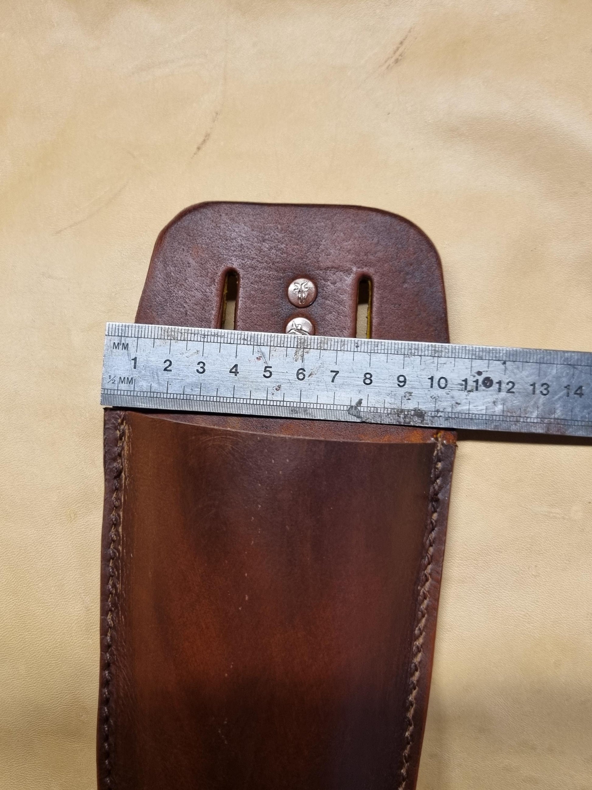 A close-up of a brown leather secateurs holster with a metal ruler laid across the top to show its width, set against a beige fabric background.