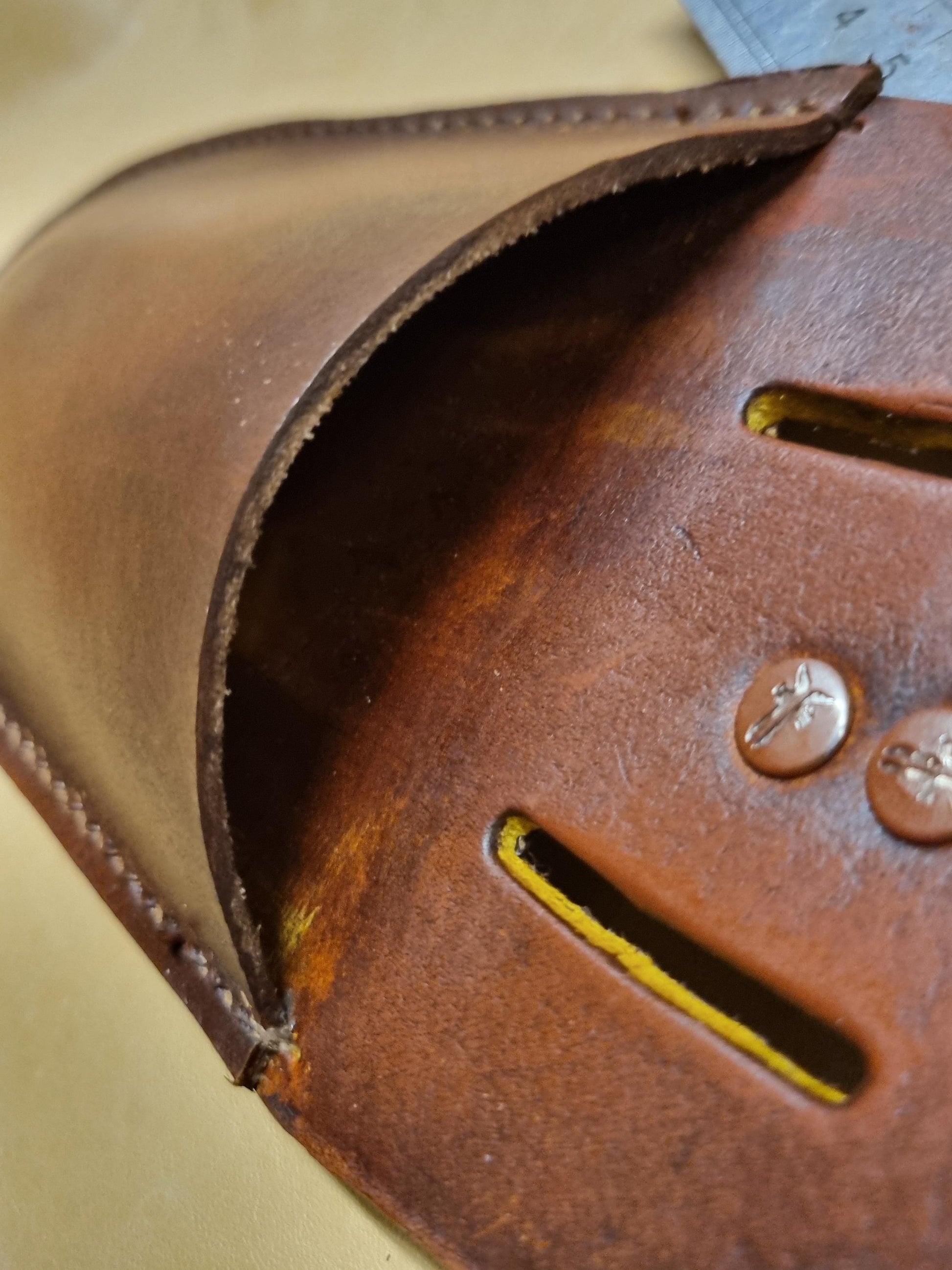 Close-up of a brown leather pouch with stitched edges, a curved opening, and two metal rivets on a beige background.