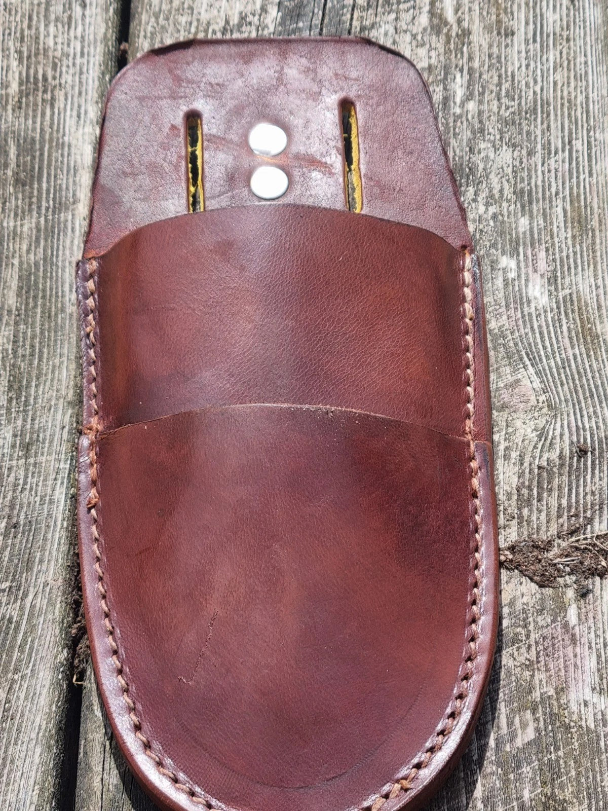 A brown leather secateurs sheath showing double pockets with decorative stitching and two metal eyelets, photographed on weathered wooden plank