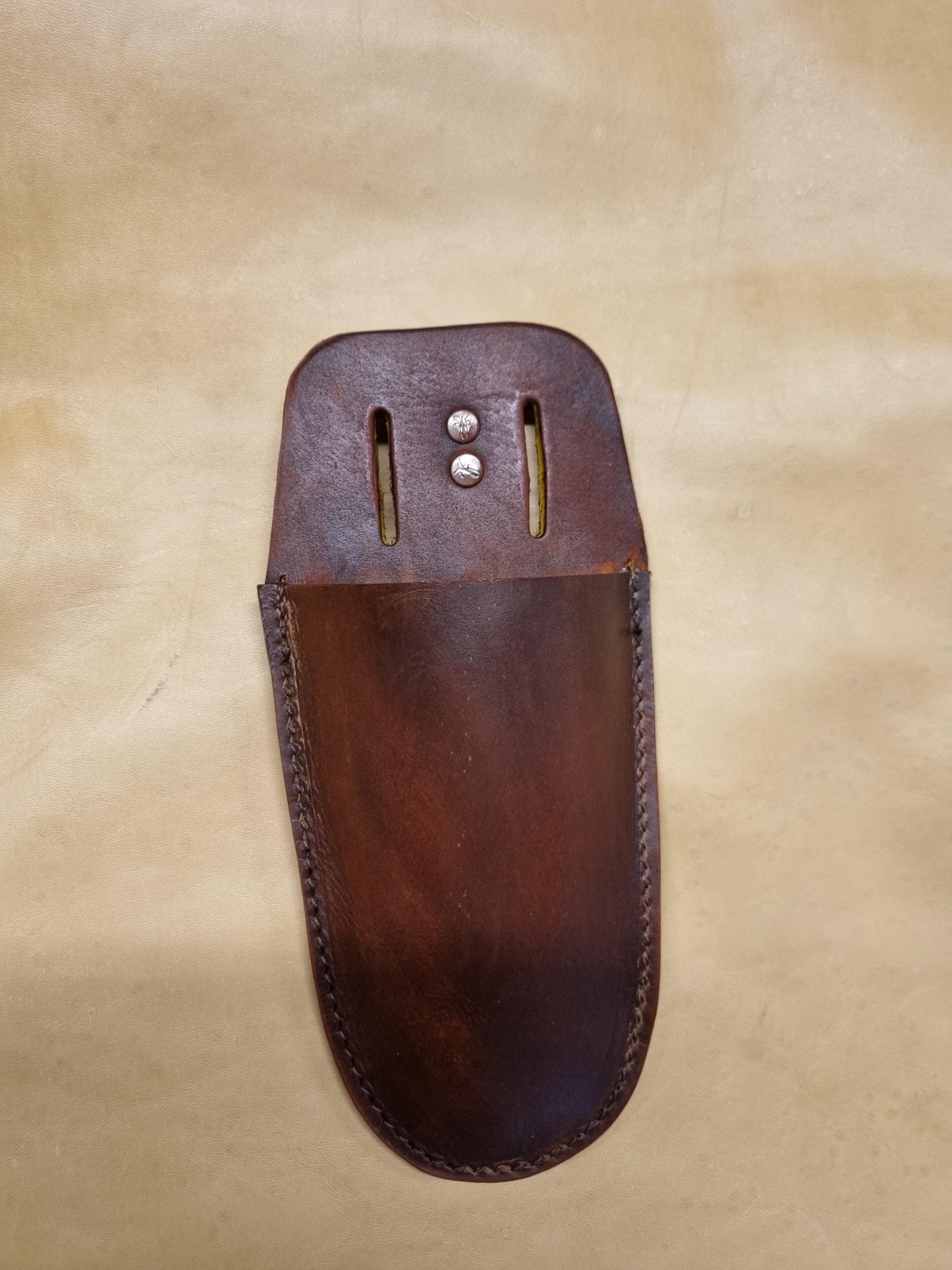 A dark brown moulded leather sheath for gardening pruners, secateurs and wire cutters showing stitch work and two metal rivets on the top flap, displayed on a textured beige background.