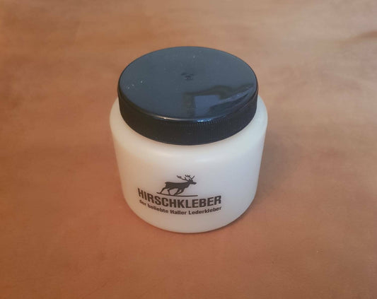 Hirschkleber paste glue tub adhesive for leather and fabric. Cream tub with black plastic lid displayed   n tan leather background