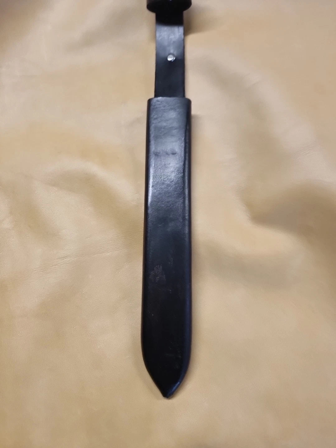 A black leather knife sheath with a matching black handle, laid vertically on a beige fabric background.
