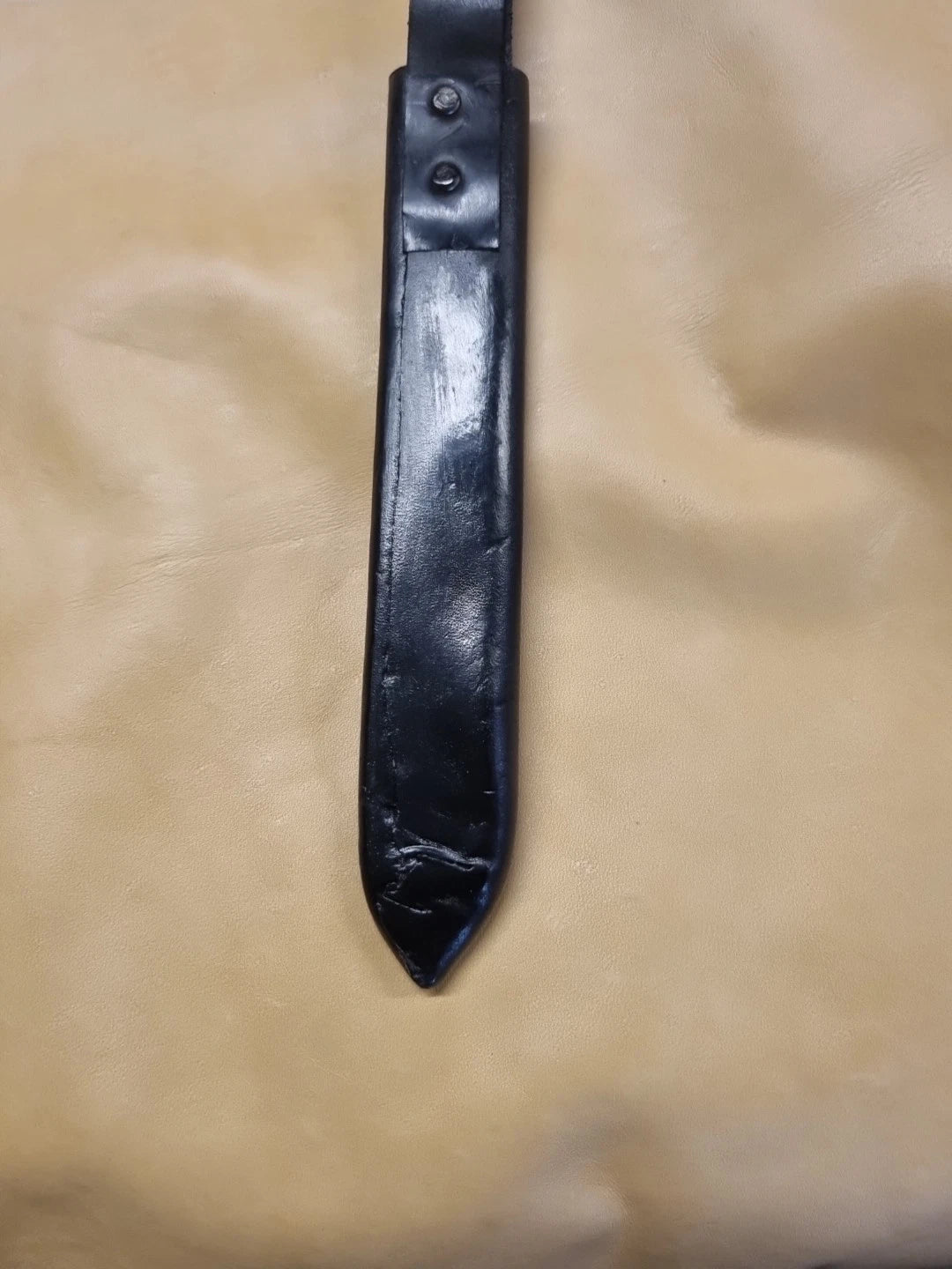 A black leather knife sheath with a pointed tip and two rivets, laid flat on a beige fabric background.