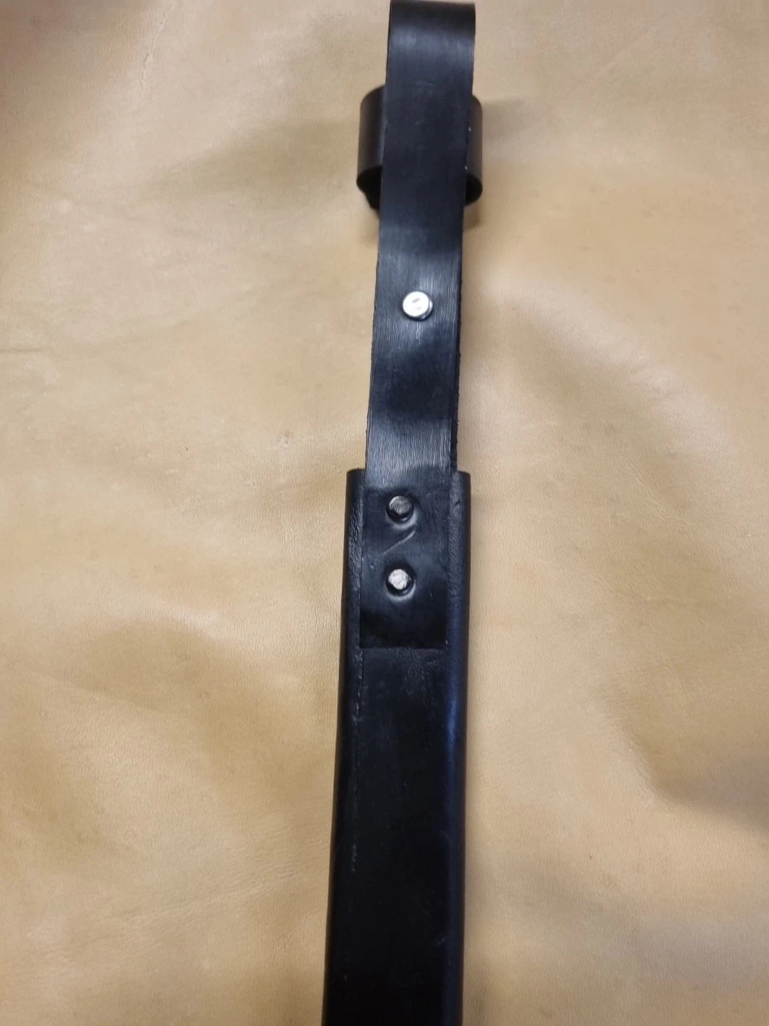 A close-up of a back of the black leather knife sheath with belt section with silver metal rivets, laid flat on a tan leather background.