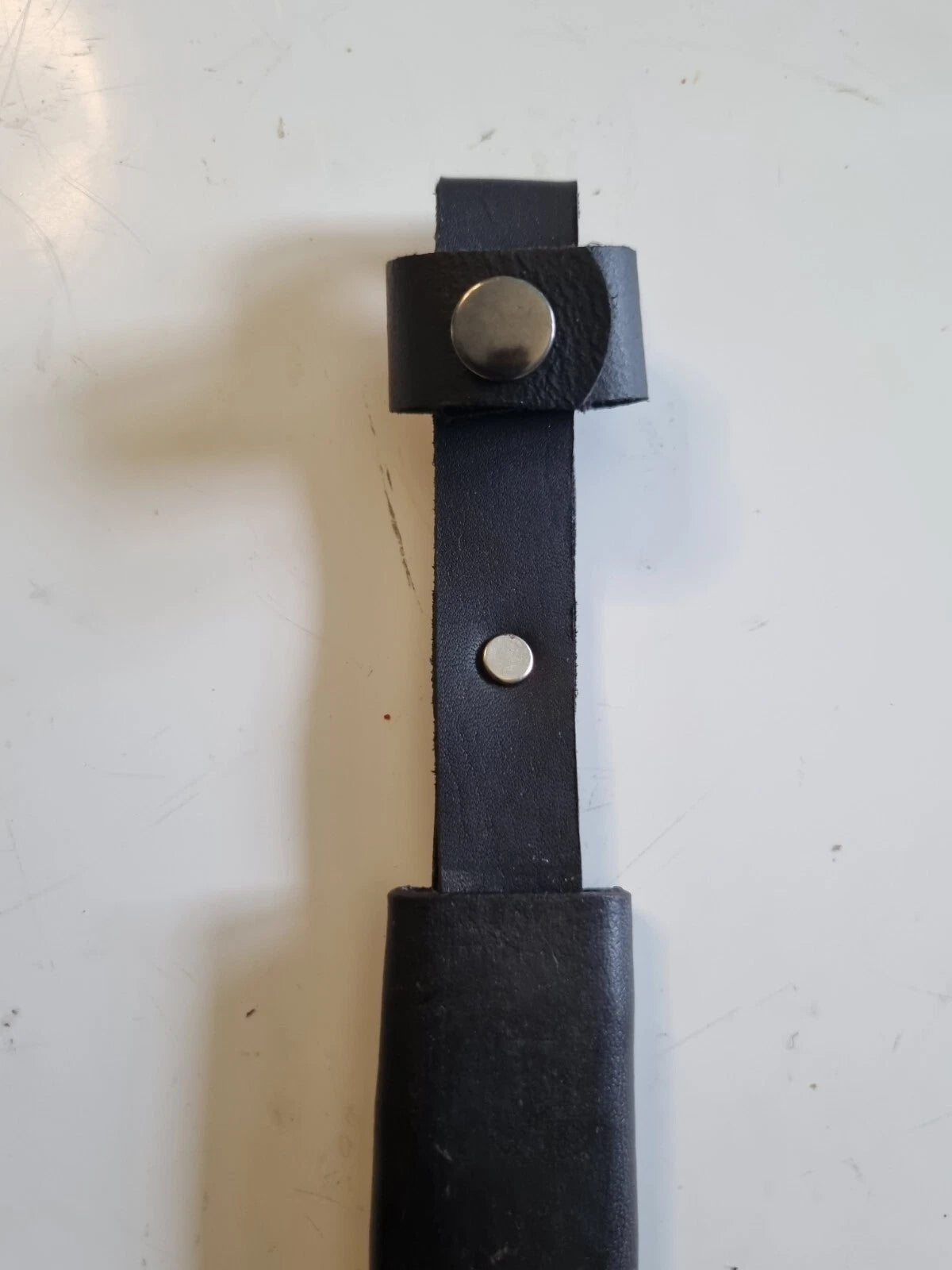 A close-up of a black leather strap or handle with two silver metal rivets, resting on a white surface.