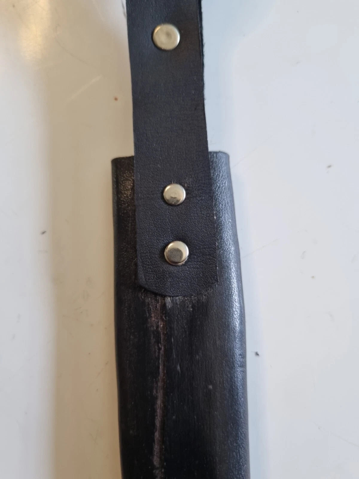 Close-up of a black leather knife sheath with three silver metal rivets, shown on a light off-white background.