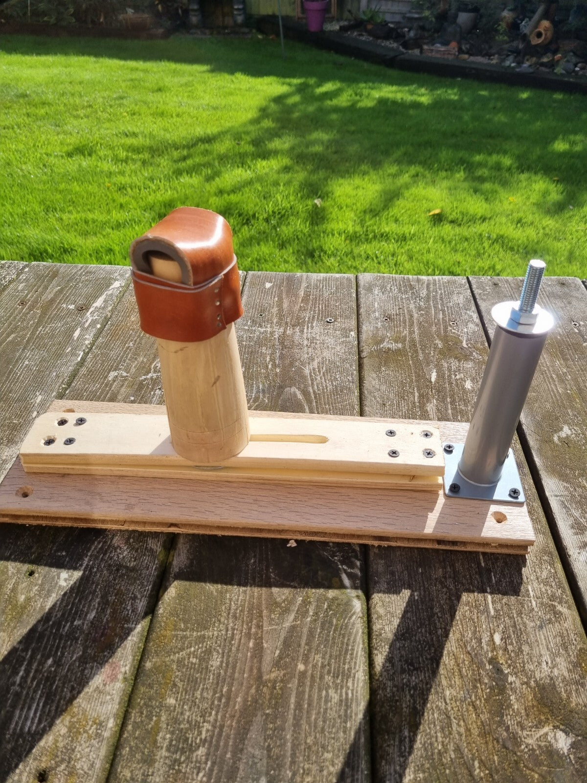 A wooden and metal shoemakers lasting stand with shoe last rest jig resting on an outdoor wooden table, with a green grass lawn in the background.