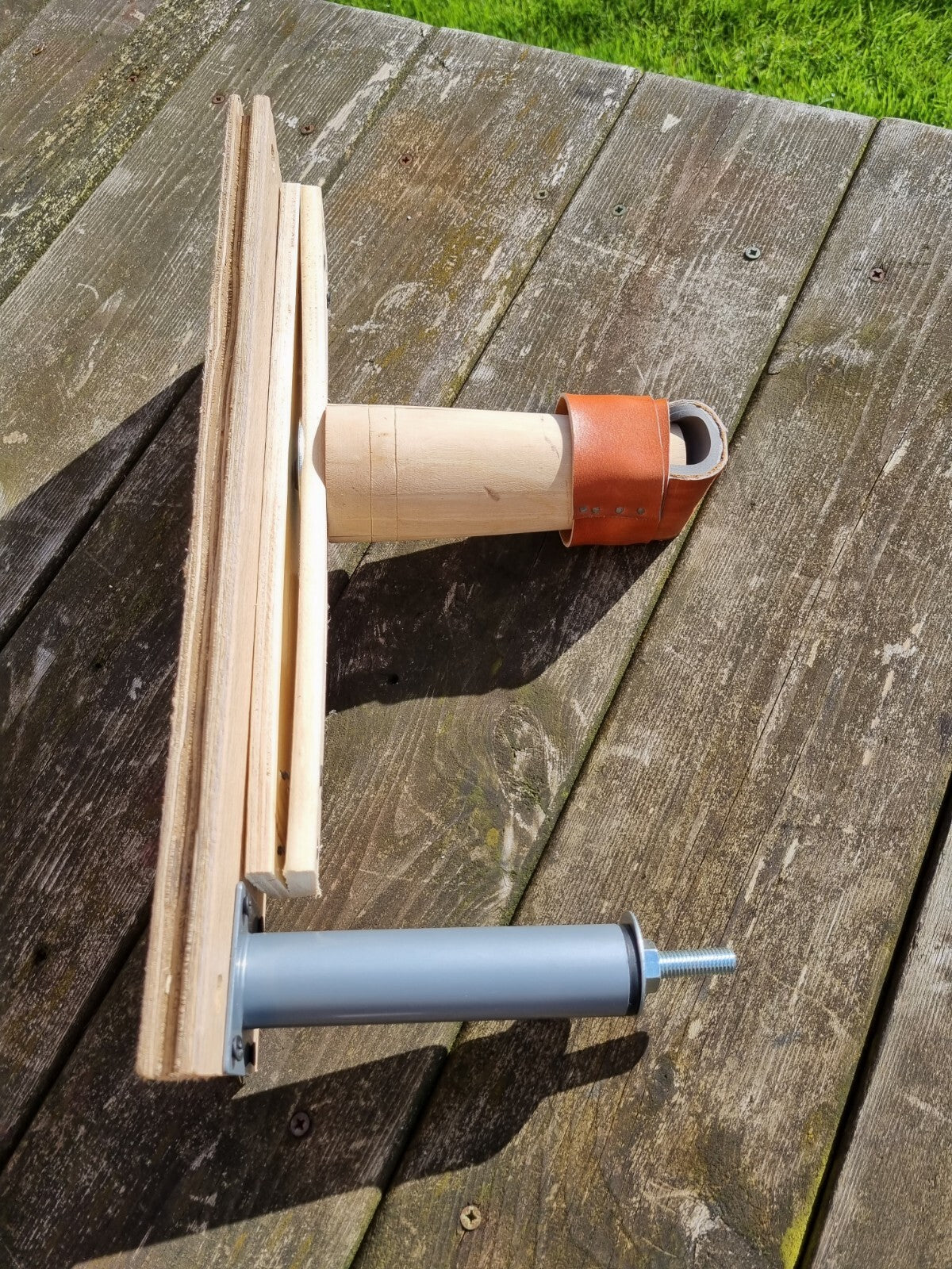 A custom handmade lasting jack stand component made of natural wood, a grey metal cylinder, and a brown leather covered post, resting on a weathered wooden deck with grass visible in the background.