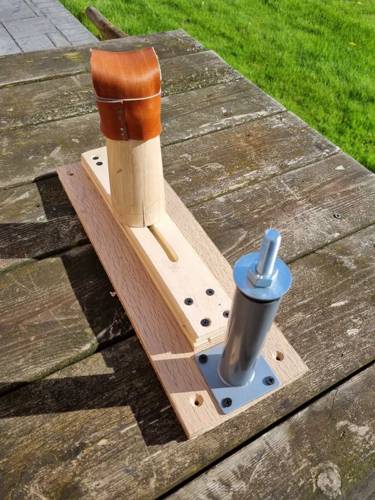 A handmade wooden and metal mechanical shoe lasting device, a custom jig, resting on a weathered wooden table with grass in the background.