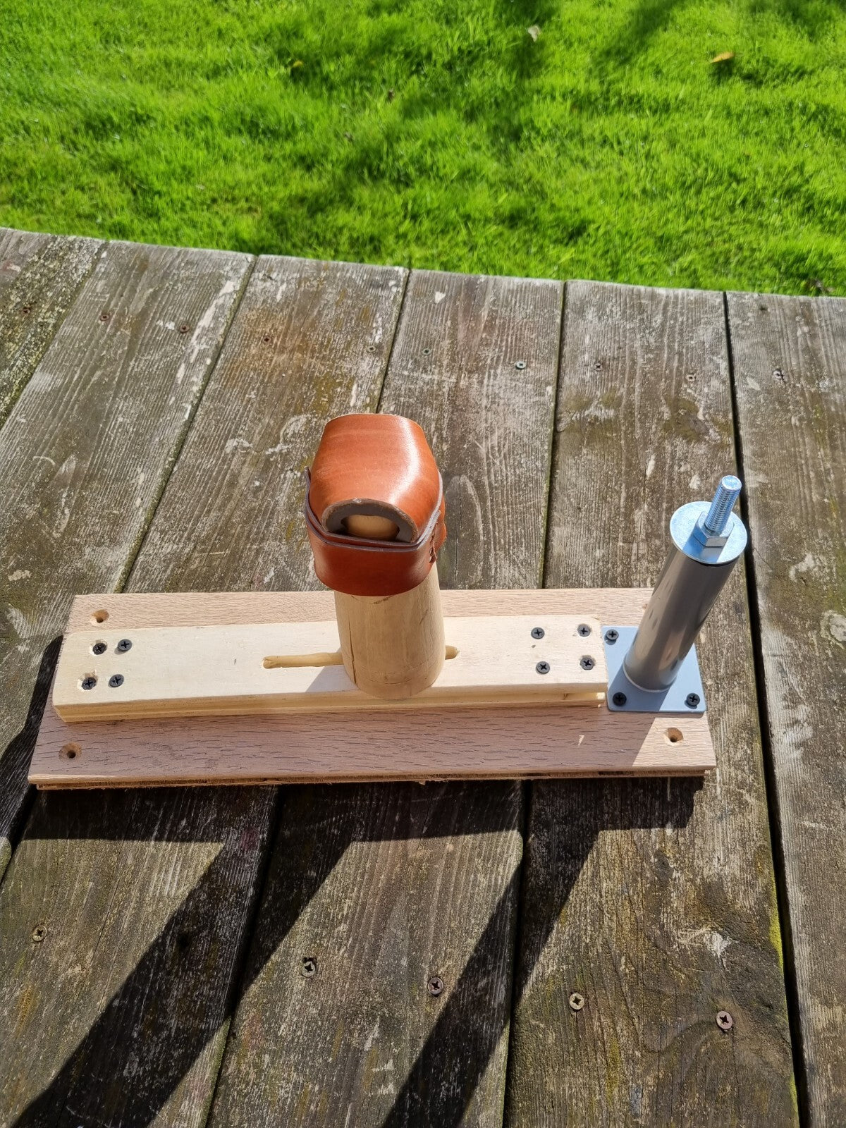 A handmade wooden and metal mechanical shoe lasting device with shoe last rest jig resting on weathered wooden deck planks with green grass in the background.