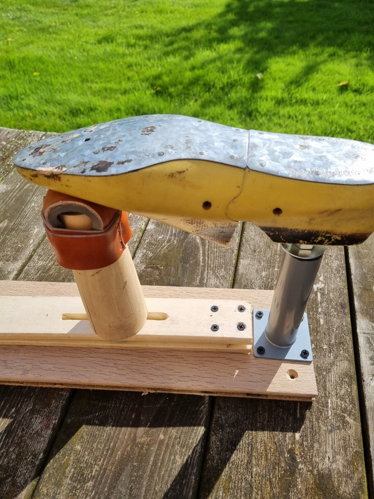 A yellow moulded plastic shoe last with silver metal foot plate mounted on a wooden base with last stand and rest, photographed outdoors on a weathered wooden deck with green grass in the background.