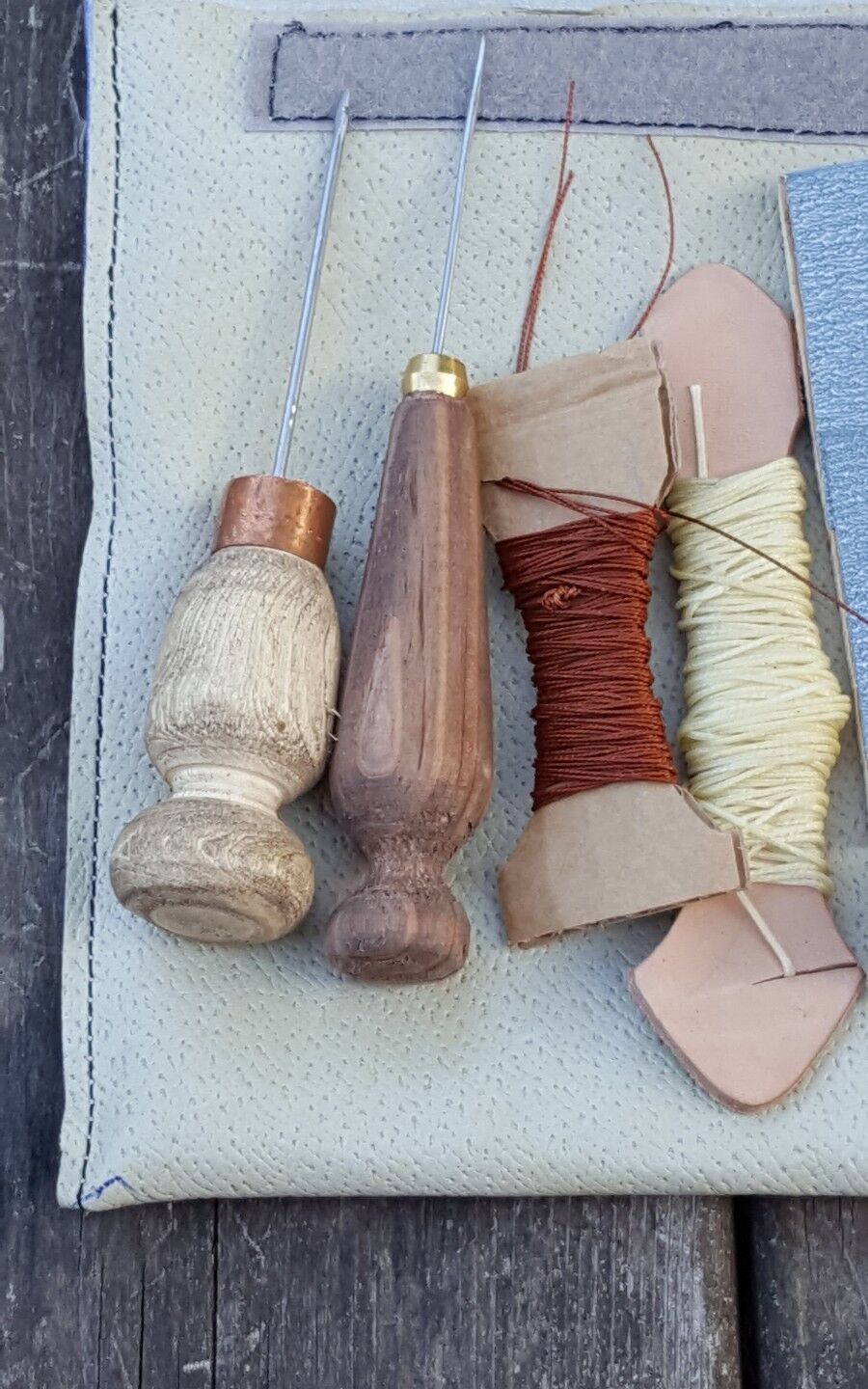 An assortment of hand sewing tools laid out on a surface, including two straight awls with wooden handles and metal hooks, a spool of brown patching thread, and a piece of fine emery paper.