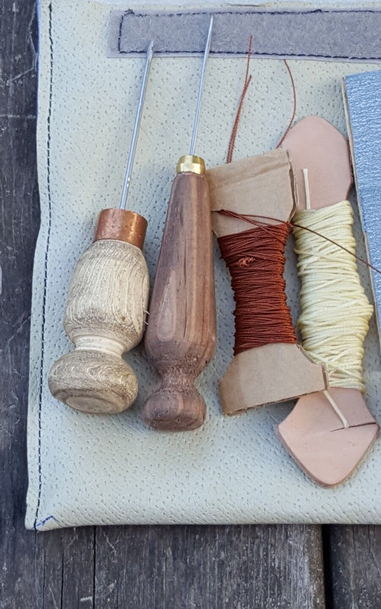 An assortment of hand sewing tools laid out on a surface, including two straight awls with wooden handles and metal hooks, a spool of brown patching thread, and a piece of fine emery paper.