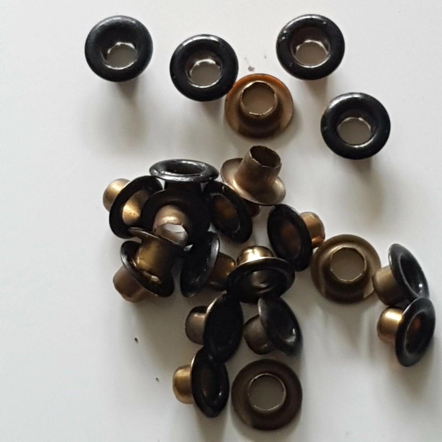 A pile of metal grommets and eyelets in black and antique bronze finishes, arranged on a plain white background.