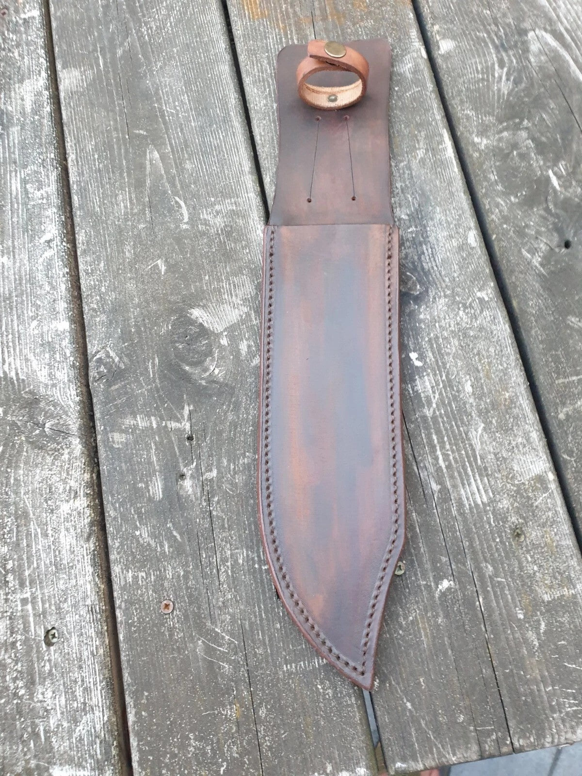 A brown leather knife sheath with a handle loop at the top, laid flat on weathered grae wooden planks.