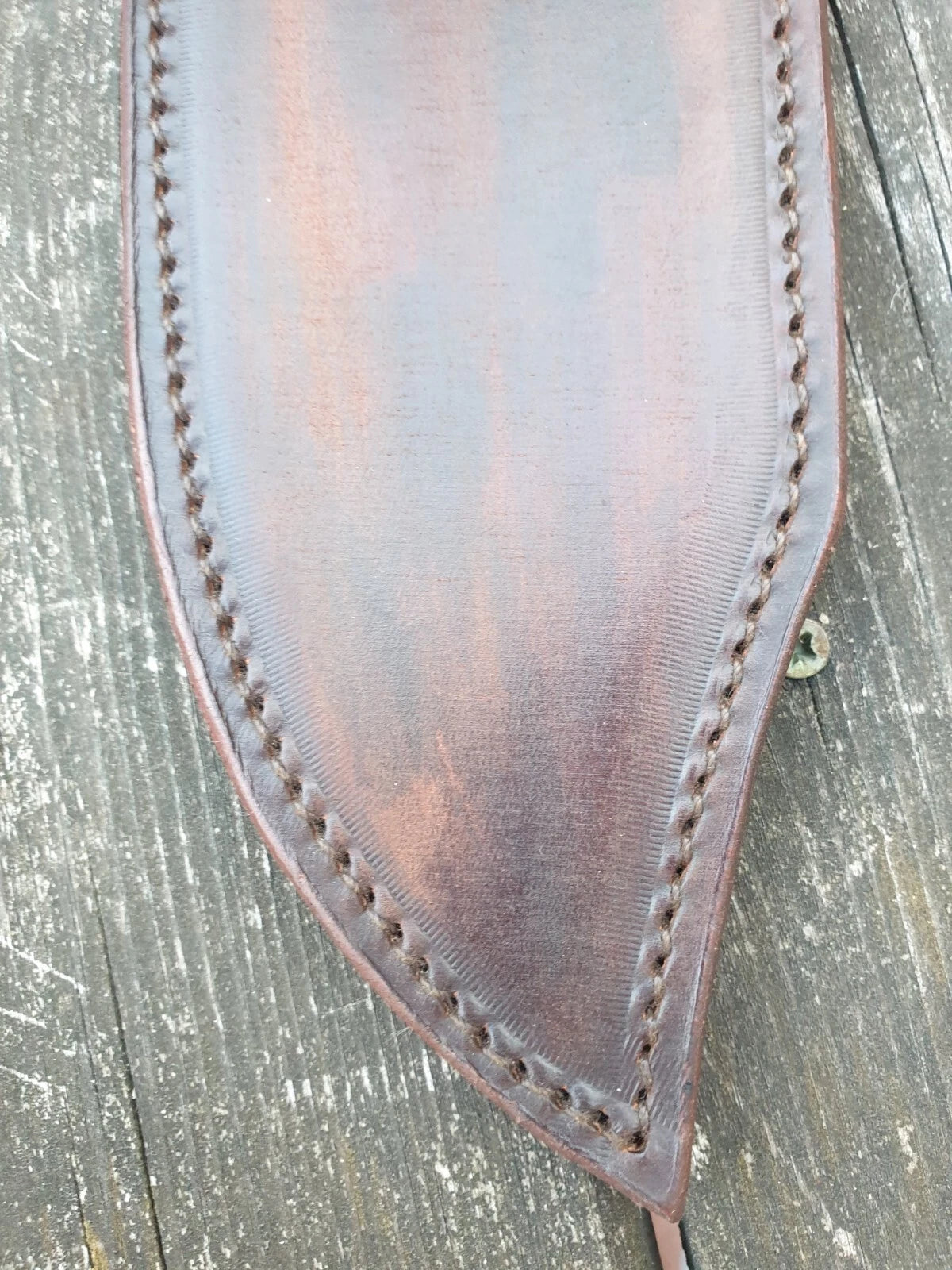 A close-up of a brown leather sheath with decorative whipstitching, laid flat on a weathered gray wooden surface.