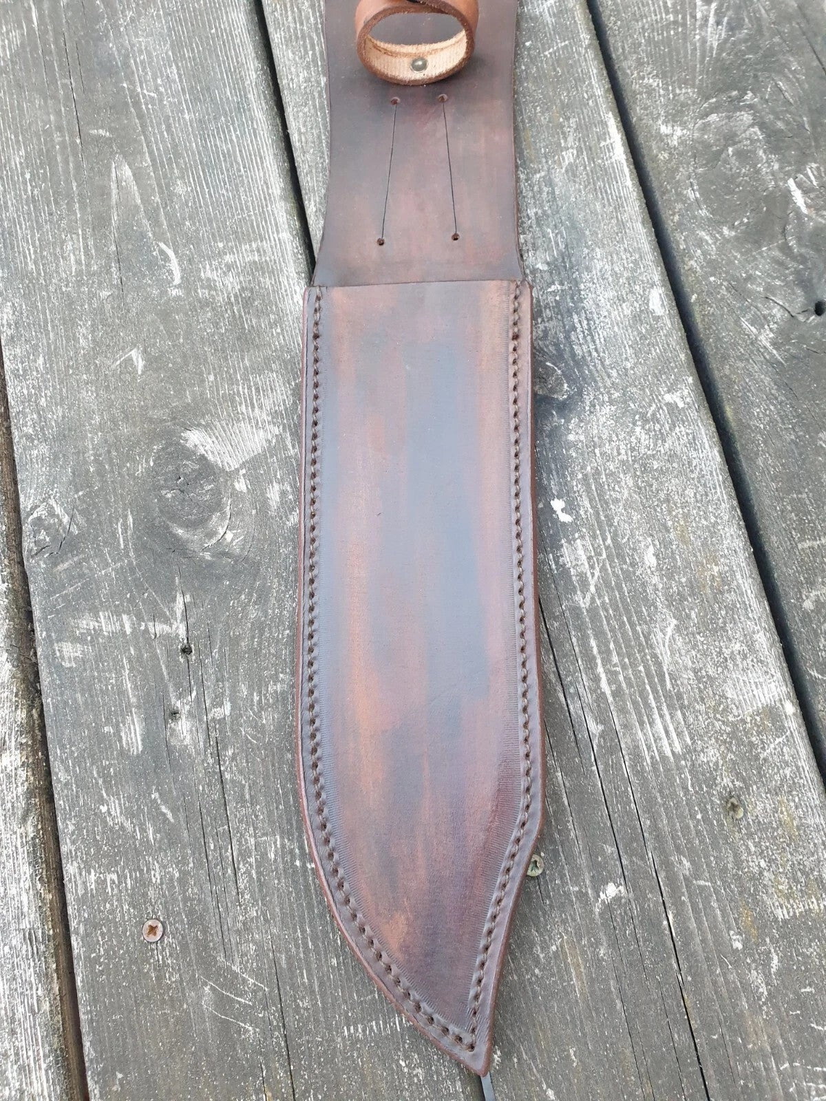 A close view of classic Bowie knife brown leather sheath with visible stitching and a handle snap loop, laid flat on weathered gray wooden planks.