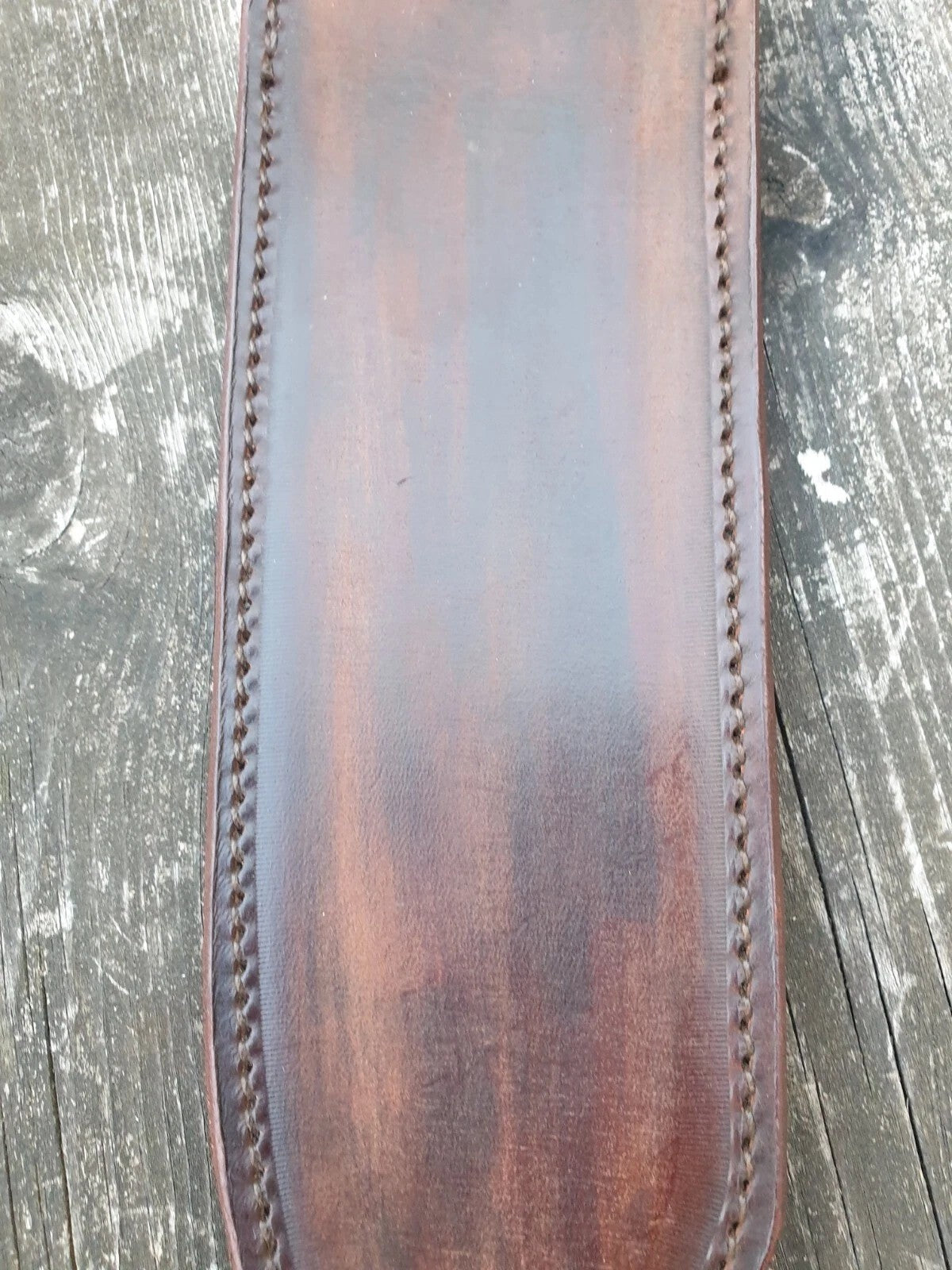 A close-up view of brown leather Bowie knife showing detailed saddle stitch work along the edges, laid flat on a weathered gray wooden surface.