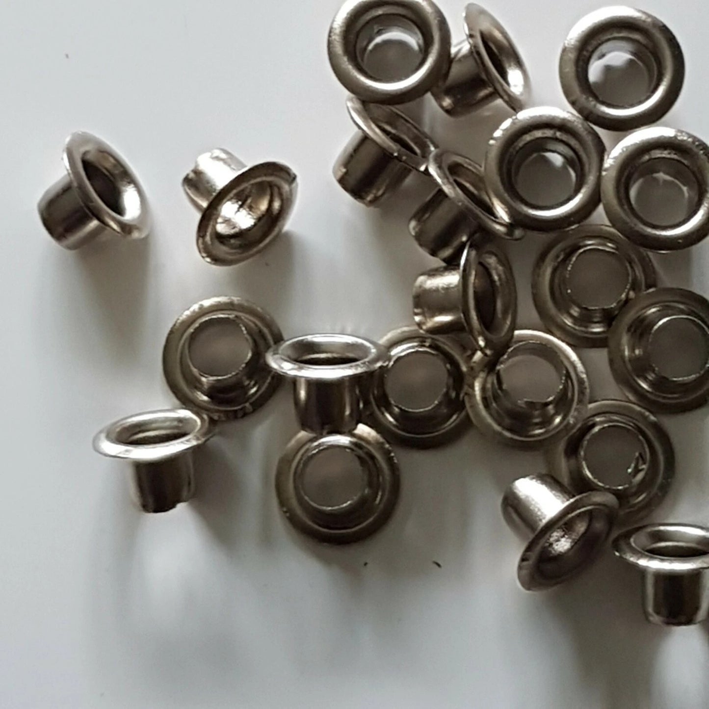 A pile of small metallic eyelets with a silver finish, scattered on a plain white surface.